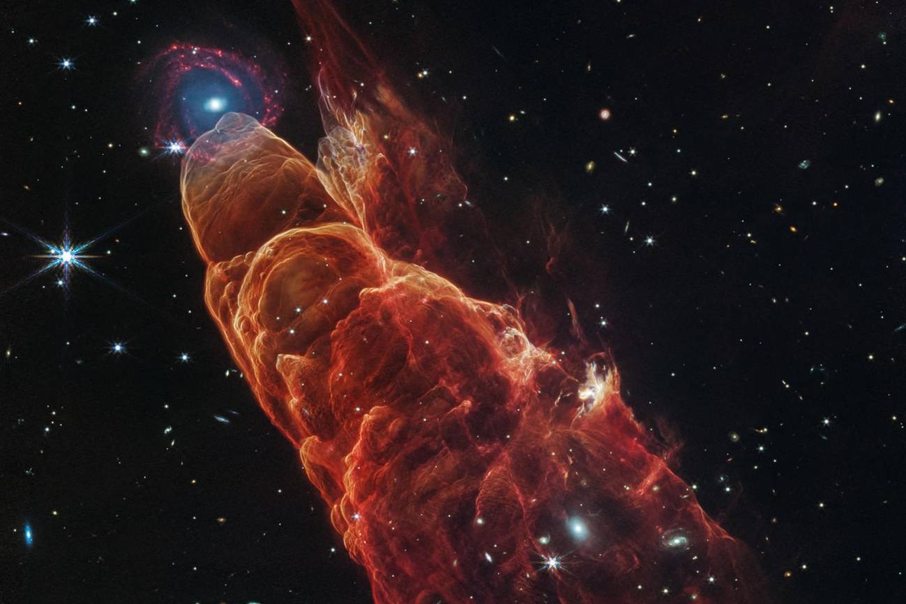 La nube de forma cónica de color rojo anaranjado corresponde a Herbig-Haro 40/50. En el extremo superior izquierdo hay una galaxia de fondo en forma de espiral con un centro azul.